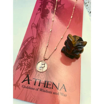 A rose gold owl-shaped pendant necklace with a matching owl figurine on a pink background, representing the Greek Goddess Athena symbol.
