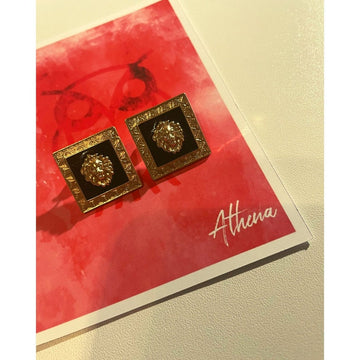 A pair of gold-colored earrings with a Medusa symbol design, displayed on a red and white card with the name 'Athena' written at the bottom.