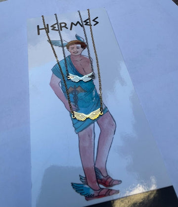 A stainless steel necklace with a winged design, displayed on a tag with an illustration of Hermes, the Greek God of commerce, wealth, and communication.