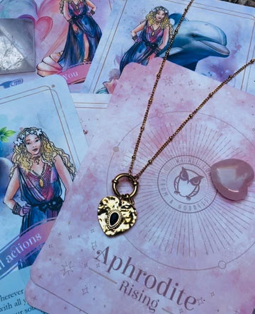 A gold-colored stainless steel necklace with a love symbol pendant, displayed on a card with an illustration of the Greek Goddess of Love, Aphrodite.
