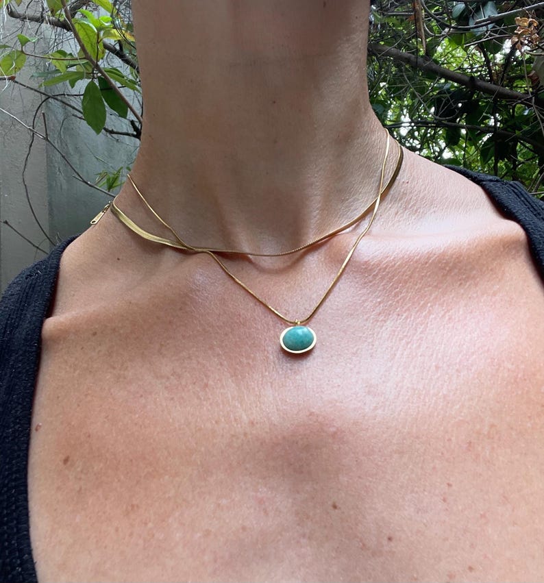 A gold-colored necklace with a teardrop-shaped amazonite pendant, presented on a pink background with text that appears to be a description or quote related to the goddess Aphrodite.