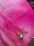A gold-colored necklace with a teardrop-shaped amazonite pendant, presented on a pink background with text that appears to be a description or quote related to the goddess Aphrodite.