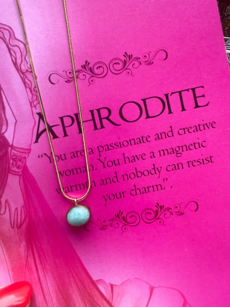 A gold-colored necklace with a teardrop-shaped amazonite pendant, presented on a pink background with text that appears to be a description or quote related to the goddess Aphrodite.