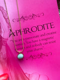 A gold-colored necklace with a teardrop-shaped amazonite pendant, presented on a pink background with text that appears to be a description or quote related to the goddess Aphrodite.