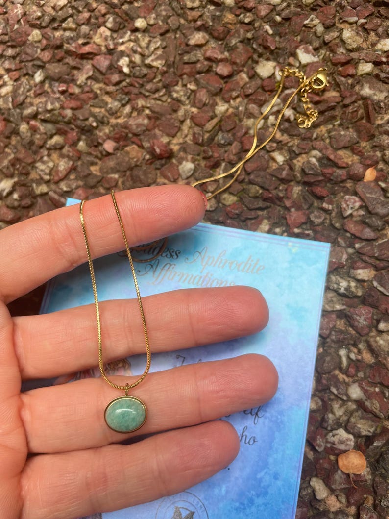 A gold-colored necklace with a teardrop-shaped amazonite pendant, presented on a pink background with text that appears to be a description or quote related to the goddess Aphrodite.