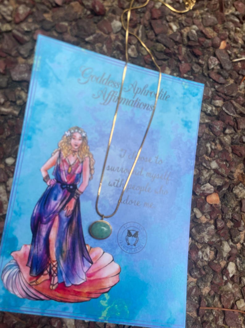 A gold-colored necklace with a teardrop-shaped amazonite pendant, presented on a pink background with text that appears to be a description or quote related to the goddess Aphrodite.