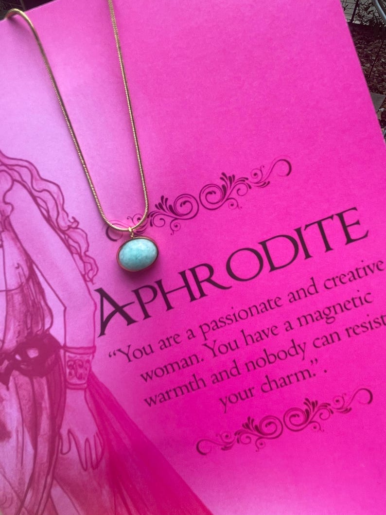 A gold-colored necklace with a teardrop-shaped amazonite pendant, presented on a pink background with text that appears to be a description or quote related to the goddess Aphrodite.