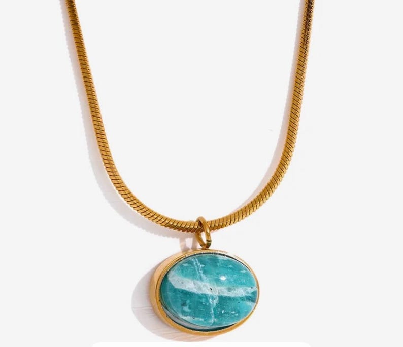 A gold-colored necklace with a teardrop-shaped amazonite pendant, presented on a pink background with text that appears to be a description or quote related to the goddess Aphrodite.