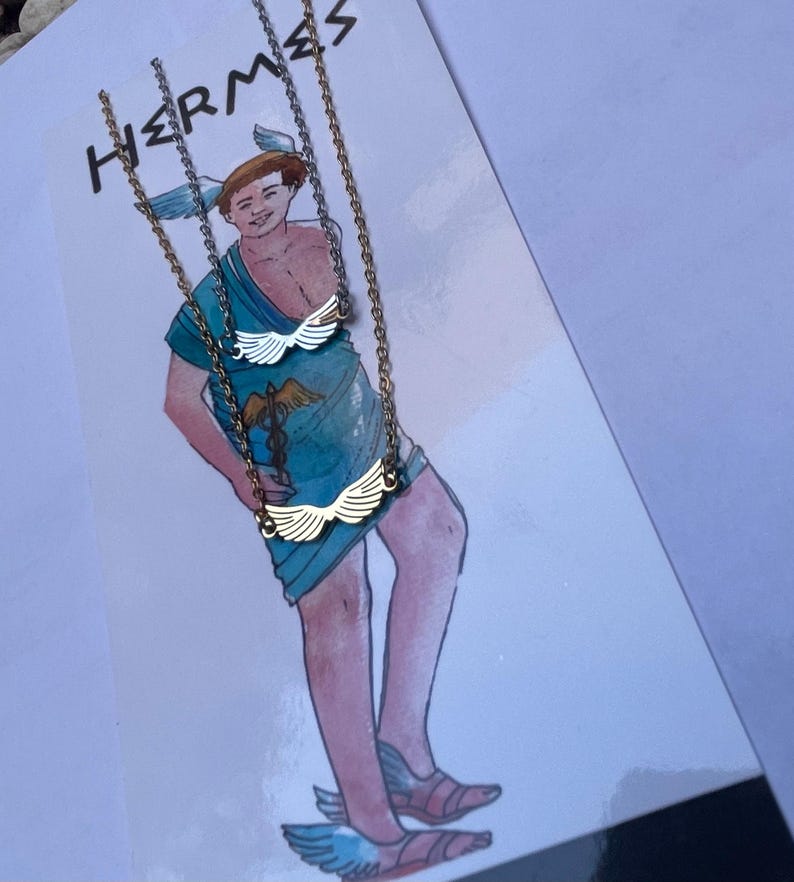 A stainless steel necklace with a winged design, displayed on a tag with an illustration of Hermes, the Greek God of commerce, wealth, and communication.