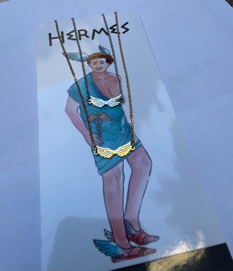 A stainless steel necklace with a winged design, displayed on a tag with an illustration of Hermes, the Greek God of commerce, wealth, and communication.