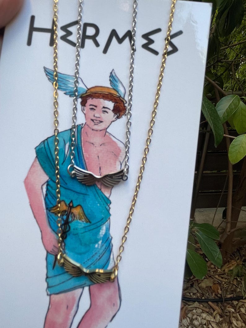 A stainless steel necklace with a winged design, displayed on a tag with an illustration of Hermes, the Greek God of commerce, wealth, and communication.