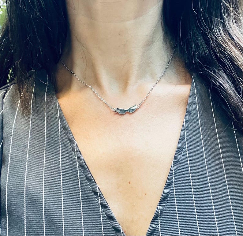 A stainless steel necklace with a winged design, displayed on a tag with an illustration of Hermes, the Greek God of commerce, wealth, and communication.