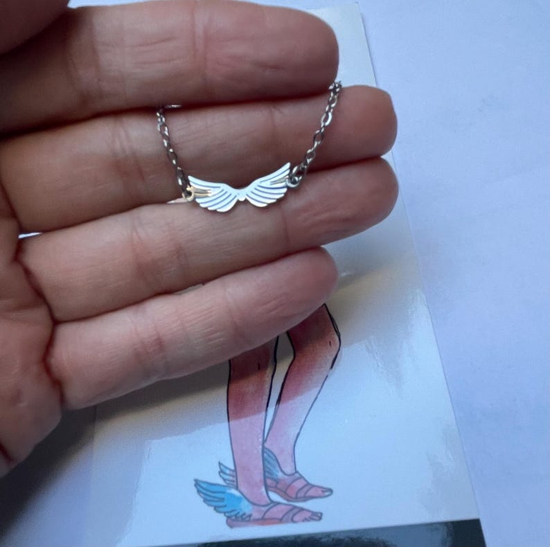 A stainless steel necklace with a winged design, displayed on a tag with an illustration of Hermes, the Greek God of commerce, wealth, and communication.