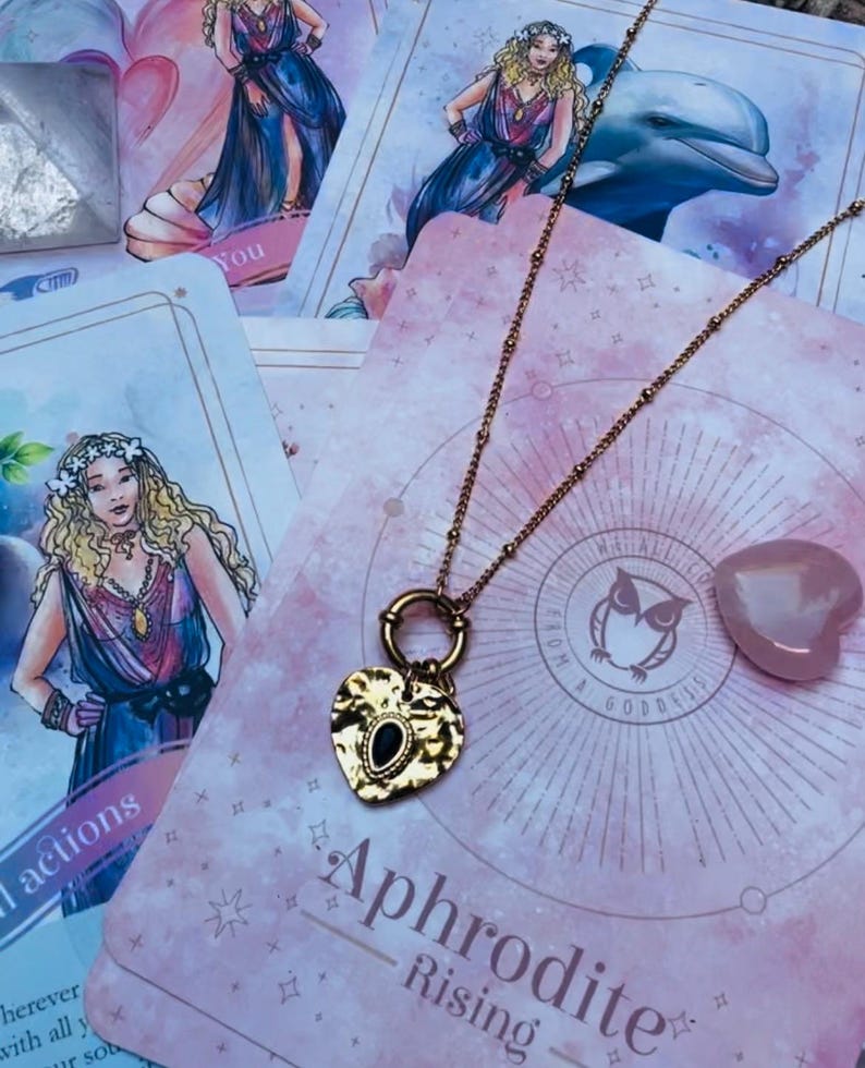 A gold-colored stainless steel necklace with a love symbol pendant, displayed on a card with an illustration of the Greek Goddess of Love, Aphrodite.