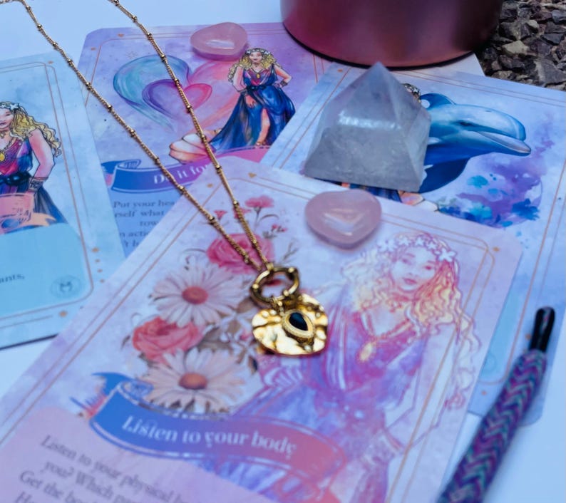 A gold-colored stainless steel necklace with a love symbol pendant, displayed on a card with an illustration of the Greek Goddess of Love, Aphrodite.