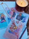 A gold-colored stainless steel necklace with a love symbol pendant, displayed on a card with an illustration of the Greek Goddess of Love, Aphrodite.
