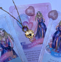 A gold-colored stainless steel necklace with a love symbol pendant, displayed on a card with an illustration of the Greek Goddess of Love, Aphrodite.