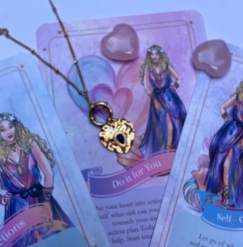 A gold-colored stainless steel necklace with a love symbol pendant, displayed on a card with an illustration of the Greek Goddess of Love, Aphrodite.