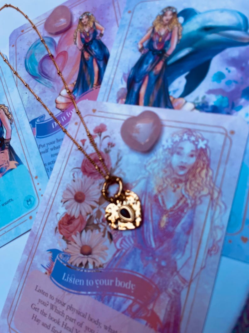 A gold-colored stainless steel necklace with a love symbol pendant, displayed on a card with an illustration of the Greek Goddess of Love, Aphrodite.