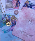 A gold-colored stainless steel necklace with a love symbol pendant, displayed on a card with an illustration of the Greek Goddess of Love, Aphrodite.
