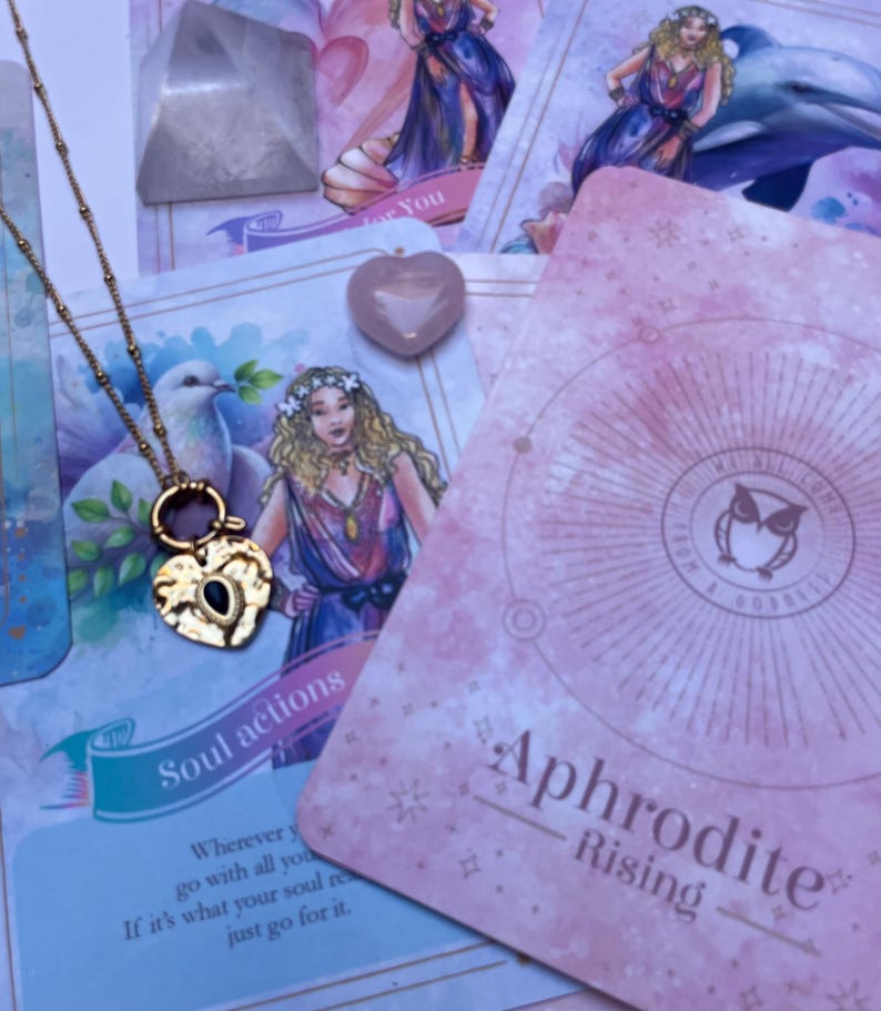 A gold-colored stainless steel necklace with a love symbol pendant, displayed on a card with an illustration of the Greek Goddess of Love, Aphrodite.