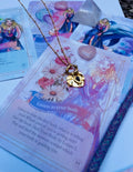 A gold-colored stainless steel necklace with a love symbol pendant, displayed on a card with an illustration of the Greek Goddess of Love, Aphrodite.