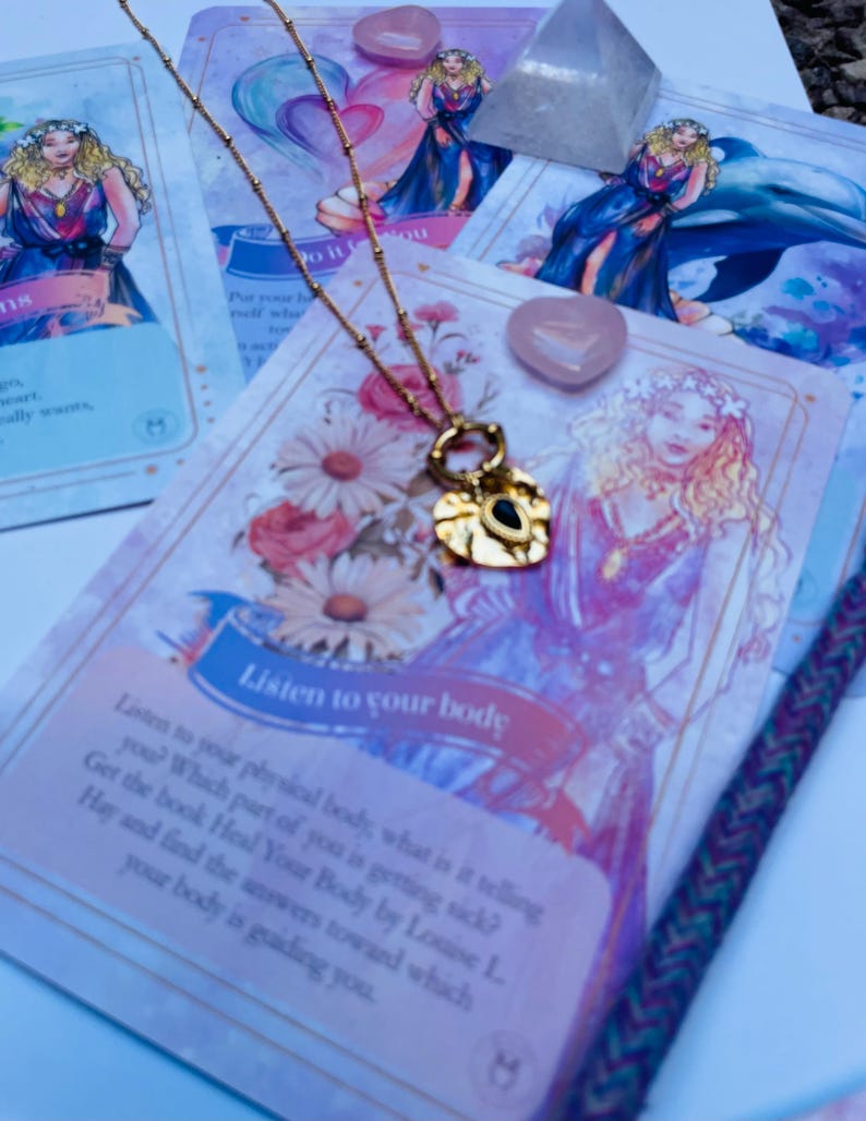 A gold-colored stainless steel necklace with a love symbol pendant, displayed on a card with an illustration of the Greek Goddess of Love, Aphrodite.