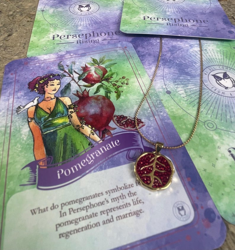 A gold-colored necklace with a pomegranate-shaped pendant, held in a person's hand, with a background featuring a book or card with 'Persphone Rising' text and celestial imagery.