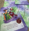 A gold-colored necklace with a pomegranate-shaped pendant, held in a person's hand, with a background featuring a book or card with 'Persphone Rising' text and celestial imagery.