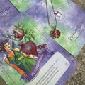 A gold-colored necklace with a pomegranate-shaped pendant, held in a person's hand, with a background featuring a book or card with 'Persphone Rising' text and celestial imagery.