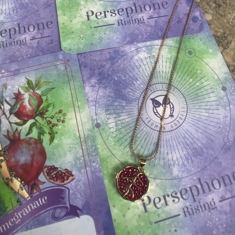 A gold-colored necklace with a pomegranate-shaped pendant, held in a person's hand, with a background featuring a book or card with 'Persphone Rising' text and celestial imagery.