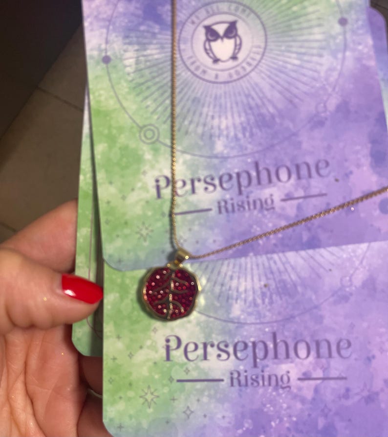 A gold-colored necklace with a pomegranate-shaped pendant, held in a person's hand, with a background featuring a book or card with 'Persphone Rising' text and celestial imagery.