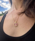 A gold-colored necklace with a pomegranate-shaped pendant, held in a person's hand, with a background featuring a book or card with 'Persphone Rising' text and celestial imagery.