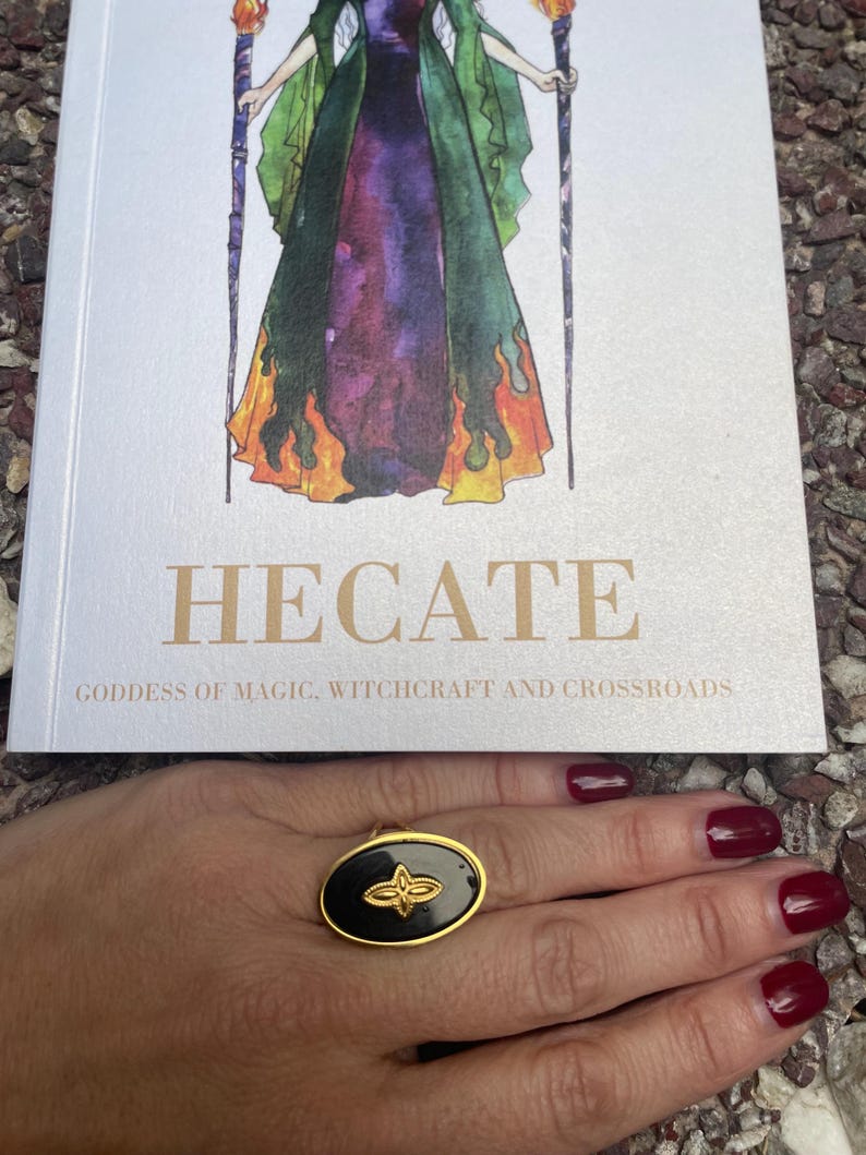 A person's hand displaying a stainless steel ring with an engraved symbol, likely representing Goddess Hekate, against a backdrop of a book or printed material.