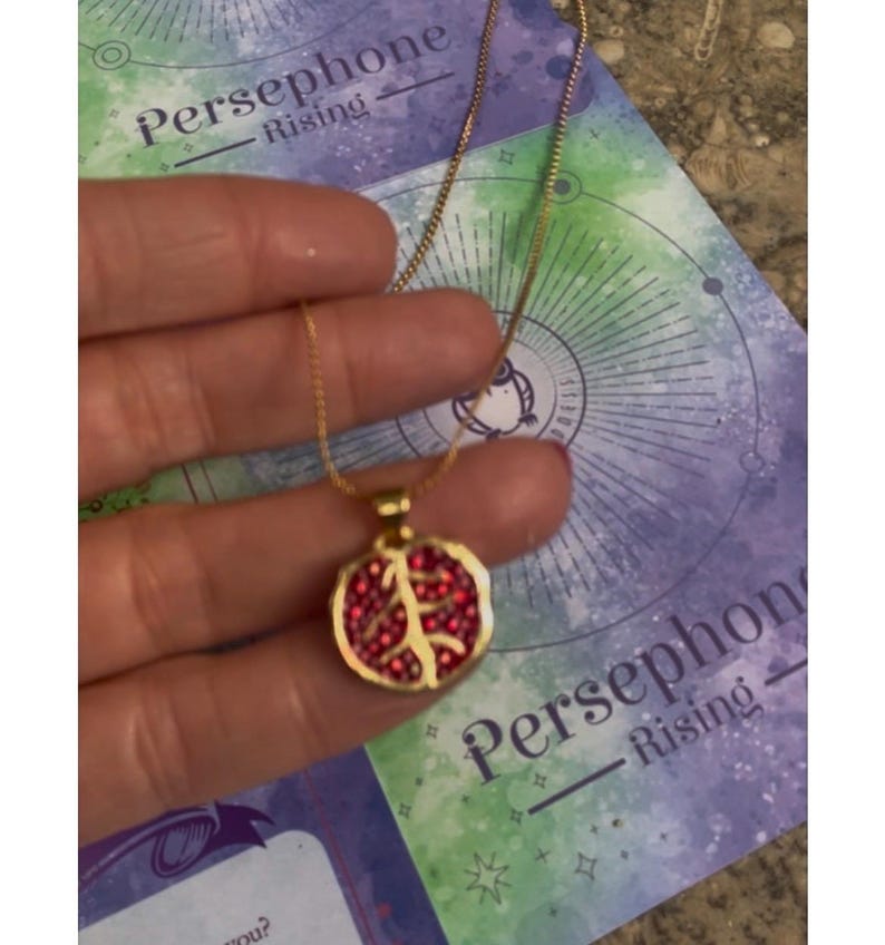 A gold-colored necklace with a pomegranate-shaped pendant, held in a person's hand, with a background featuring a book or card with 'Persphone Rising' text and celestial imagery.