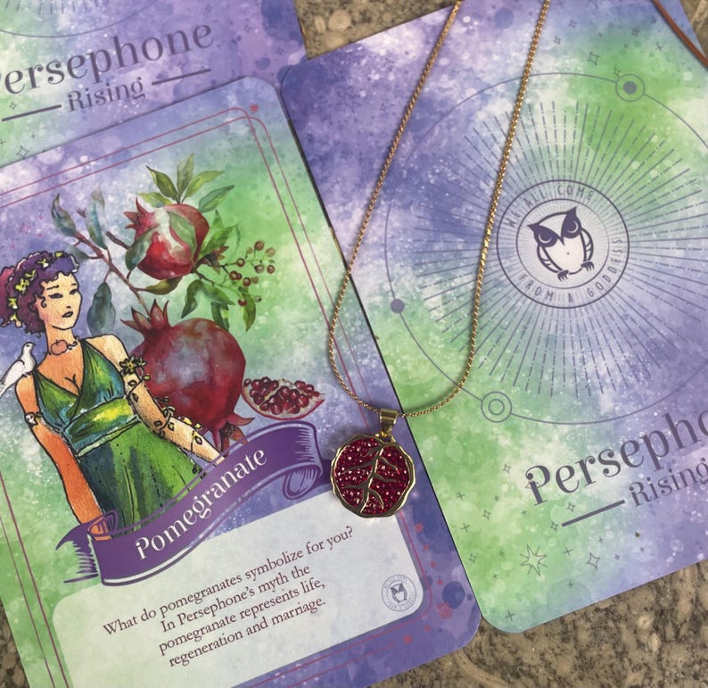 A gold-colored necklace with a pomegranate-shaped pendant, held in a person's hand, with a background featuring a book or card with 'Persphone Rising' text and celestial imagery.