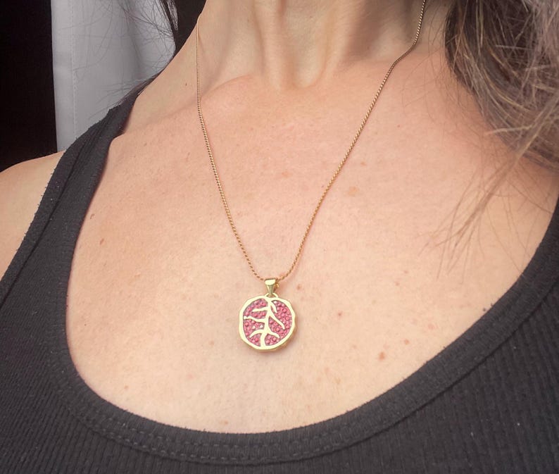 A gold-colored necklace with a pomegranate-shaped pendant, held in a person's hand, with a background featuring a book or card with 'Persphone Rising' text and celestial imagery.