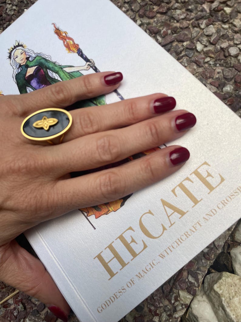 A person's hand displaying a stainless steel ring with an engraved symbol, likely representing Goddess Hekate, against a backdrop of a book or printed material.