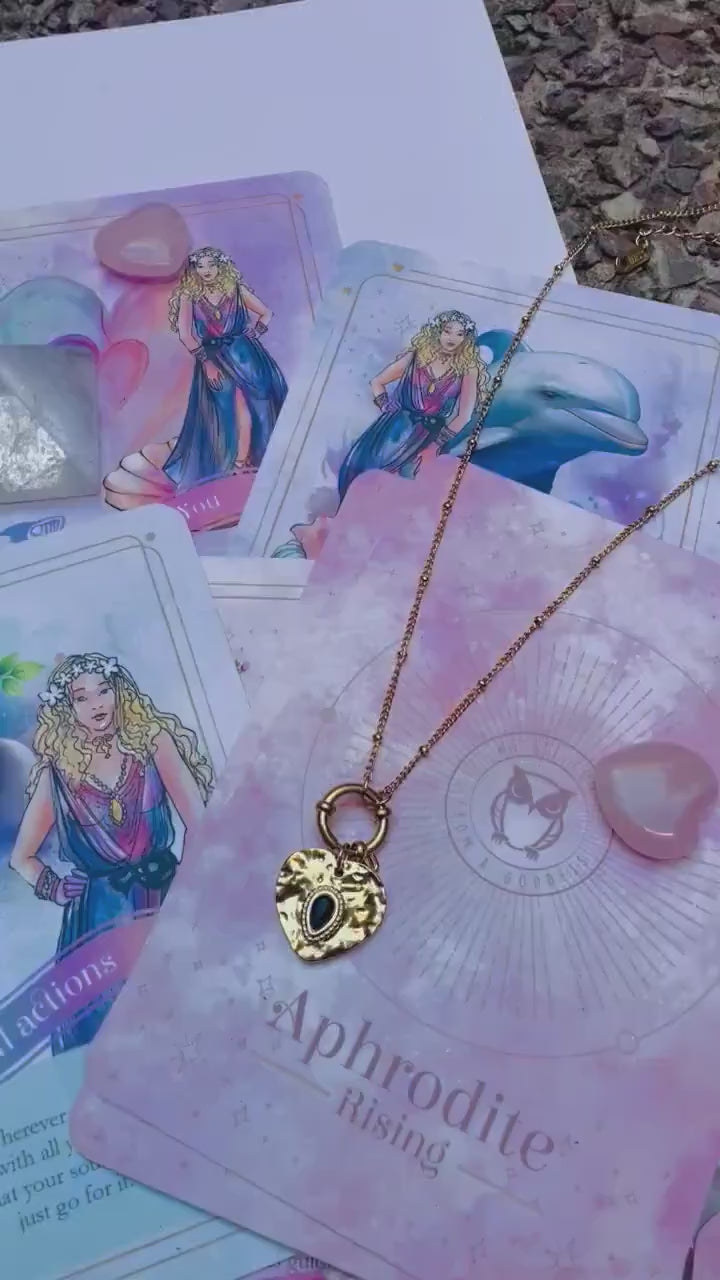 A gold-colored stainless steel necklace with a love symbol pendant, displayed on a card with an illustration of the Greek Goddess of Love, Aphrodite.